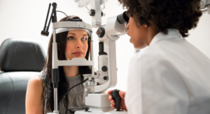 A woman with long black hair is having an eye exam with a female doctor with black short curly hair wearing a lab coat.