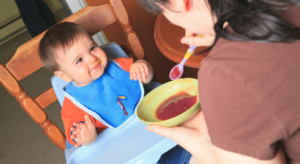 mom holding baby spoon with apple sauce to feed to child sitting in high chair