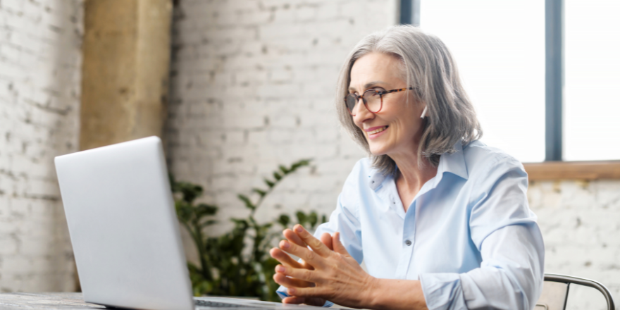 Grey-haired woman sitting at a desk in front of a laptop as if she is in a virtual meeting.