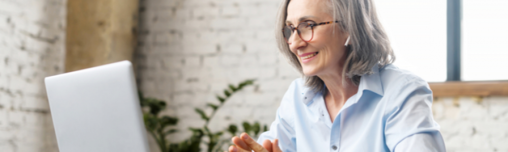 Grey-haired woman sitting at a desk in front of a laptop as if she is in a virtual meeting.