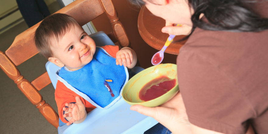 mom holding baby spoon with apple sauce to feed to child sitting in high chair