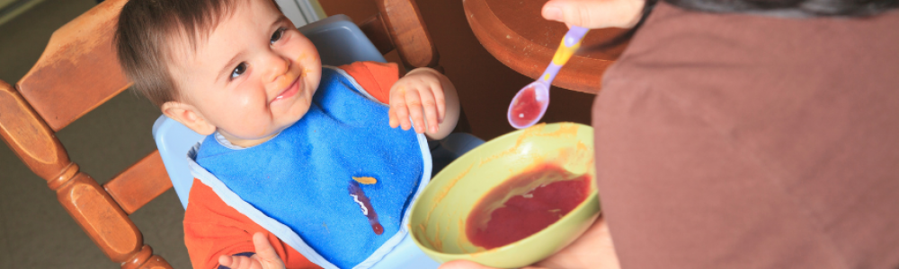 mom holding baby spoon with apple sauce to feed to child sitting in high chair